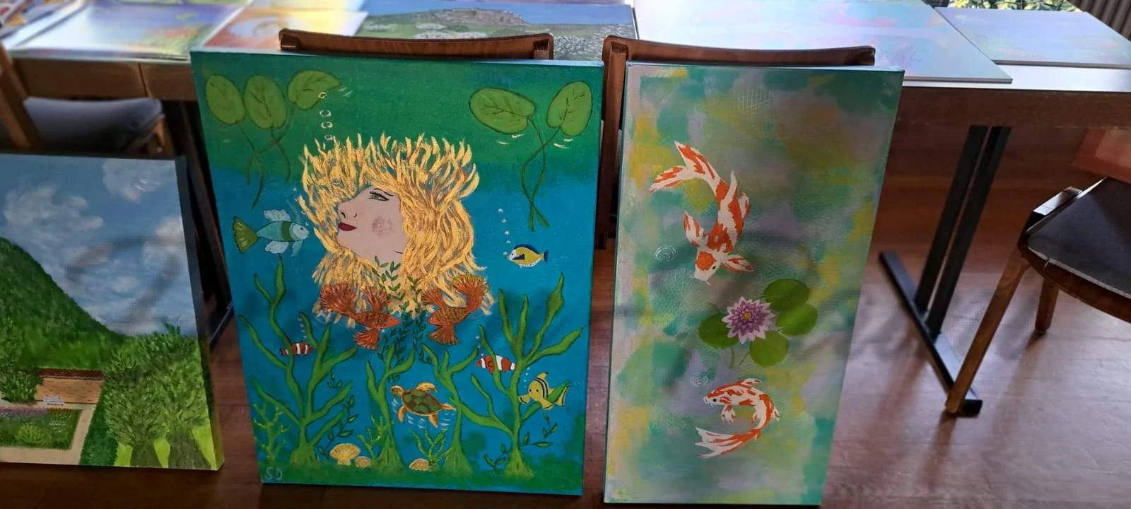 Verschiedene handgemalte Leinwandbilder auf einem Holztisch. Ein Bild zeigt ein Porträt einer Frau mit goldenem Haar, das sich in Seetang verwandelt, umgeben von Meerestieren. Das andere zeigt einen bunten Koi-Fisch in einem Teich. Im Hintergrund sind weitere Kunstwerke und Möbel zu sehen.